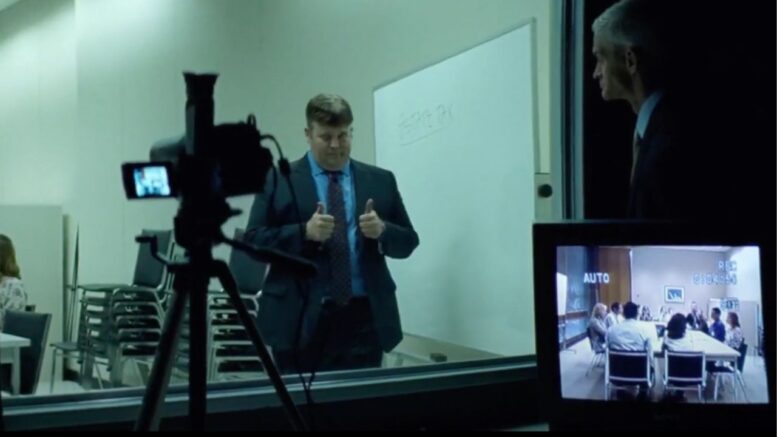 A man in a suit portraying political marketing expert and pollster Frank Luntz gives two thumbs up in front of a whiteboard during a recorded focus group session, observed from behind a glass panel.